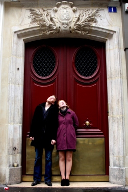 Engagement photos in Paris, France. Newlywed couple stands at door waiting for the rain to stop.